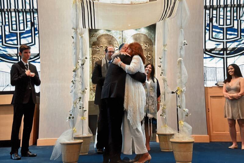 couple hugging on the bimah under the chuppah. Rabbi Miller and Cantor Azu smile from in front of the ark.